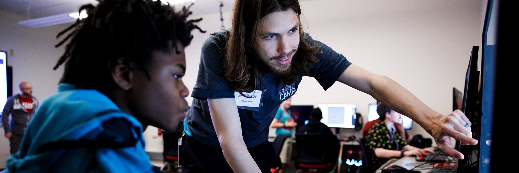 A summer camp leader points to a participant's screen as others work on computers behind them.