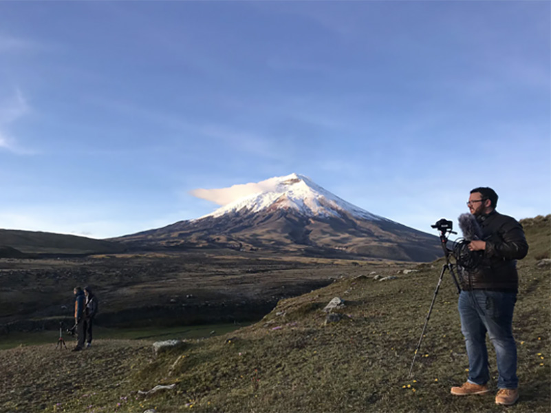 A student stands with a camera in front of a mountain in Ecuador.