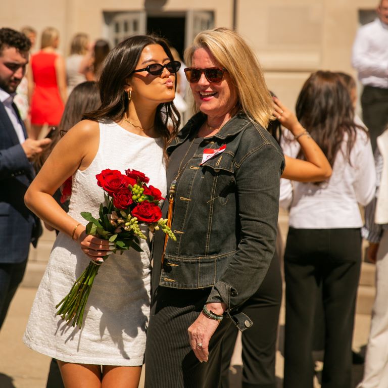 Person holding a bouquet of flowers leans to kiss a woman next to her on the cheek.